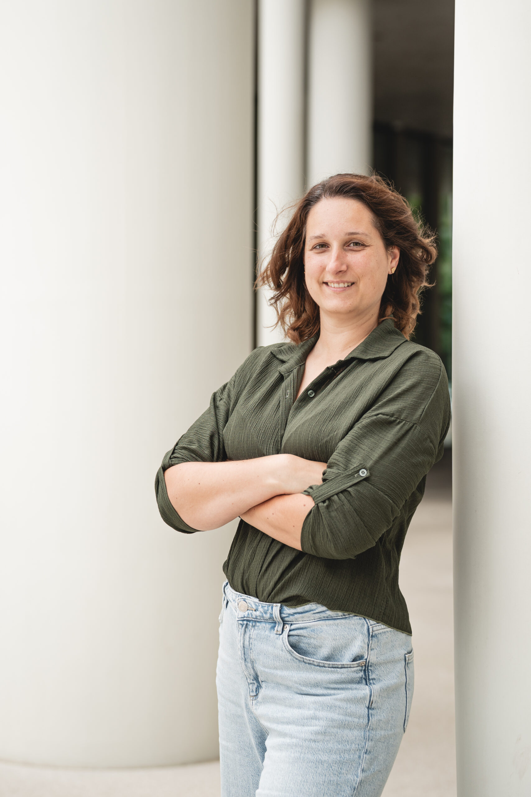 Eine Frau steht mit verschränkten Armen an einem hellen Pfeiler, trägt grüne Bluse und helle Jeans, schaut freundlich in die Kamera.
