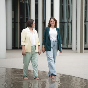 Zwei Frauen in Blazern laufen barfuß durch ein flaches Wasserbecken vor moderner Architektur.