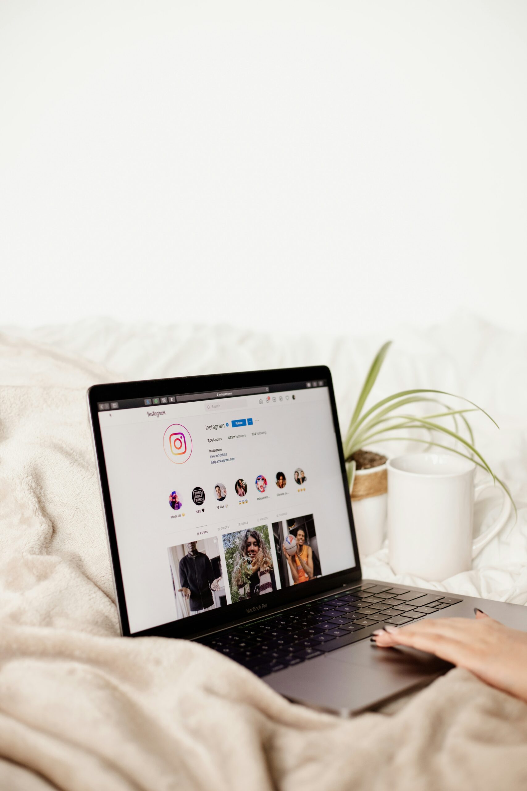Ein geöffneter Laptop zeigt die Instagram-Seite, liegend auf einer beigefarbenen Decke neben einer Pflanze und einem weißen Becher.